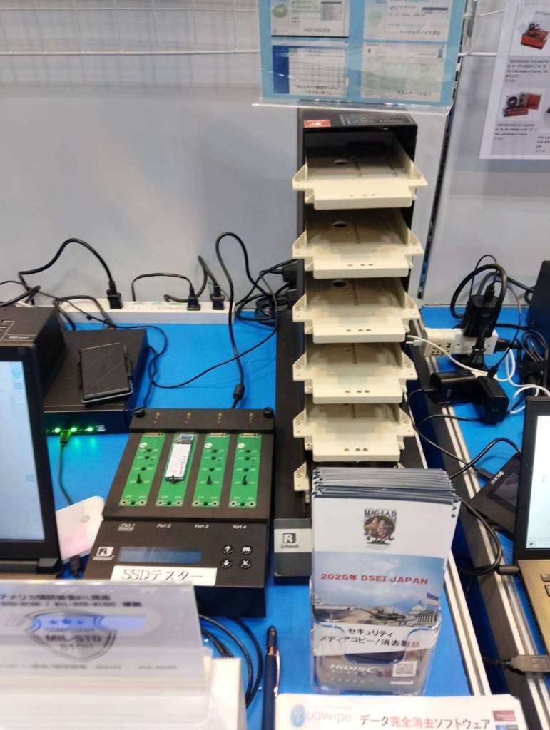 ギフトショー春2026の東京インターナショナル・ショー 共同出展ブースの青いテーブルには、SSD用のプラスチック・トレイが積み重ねられたデスクトップ・デバイスが、緑の回路基板とパンフレットの横に置かれている。.